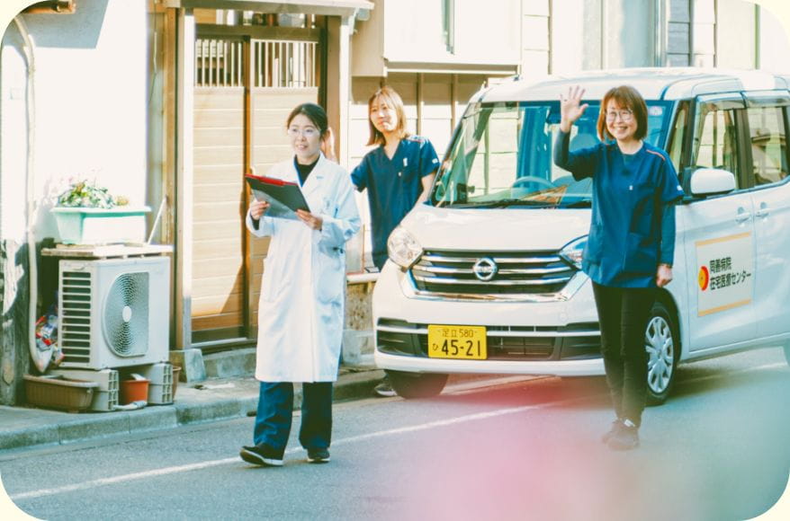 訪問診療の車の前で笑顔で手を振る医師と訪問看護スタッフ